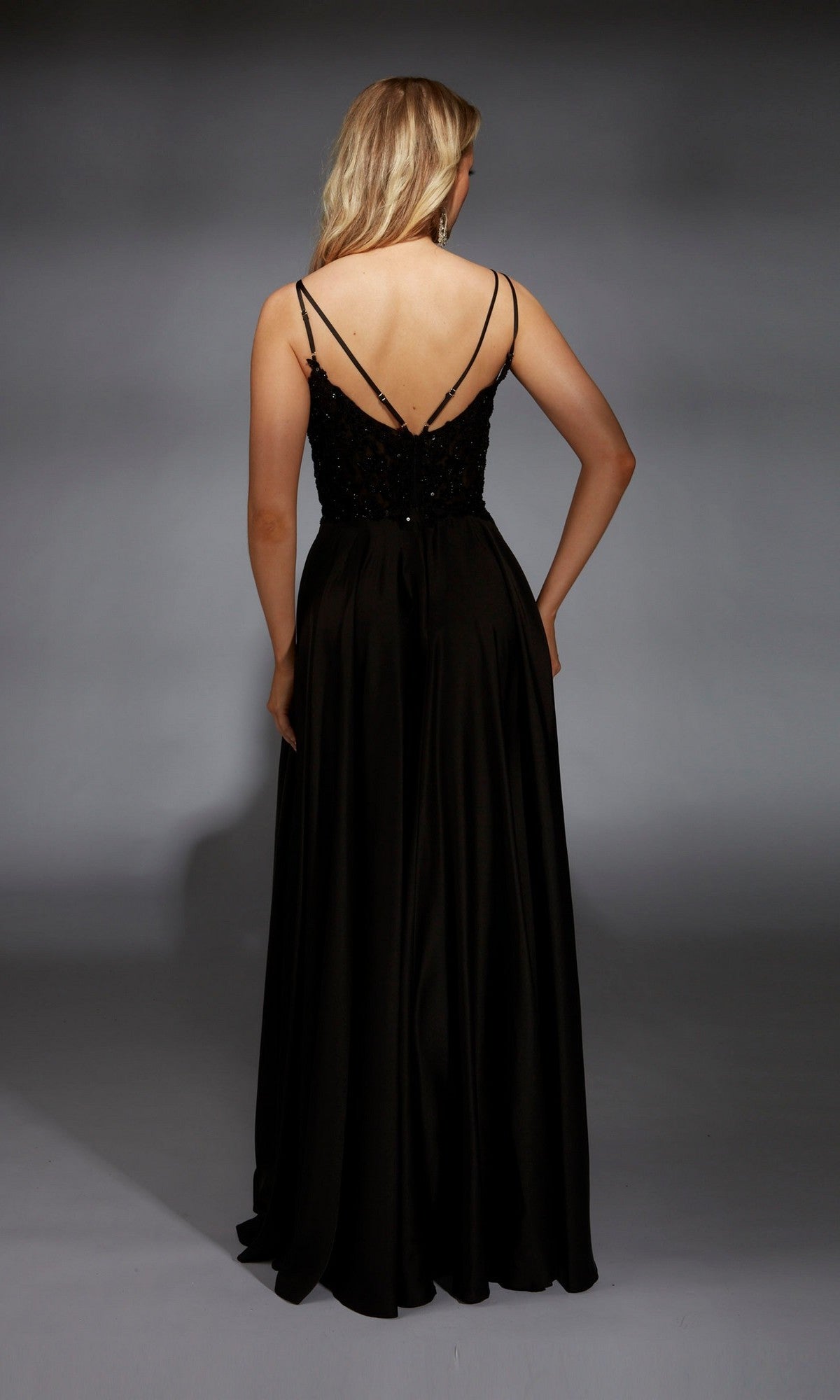 A woman stands facing away against a gray background, wearing the Aerie Couture 368 long black prom dress featuring an embellished bodice and thin straps.